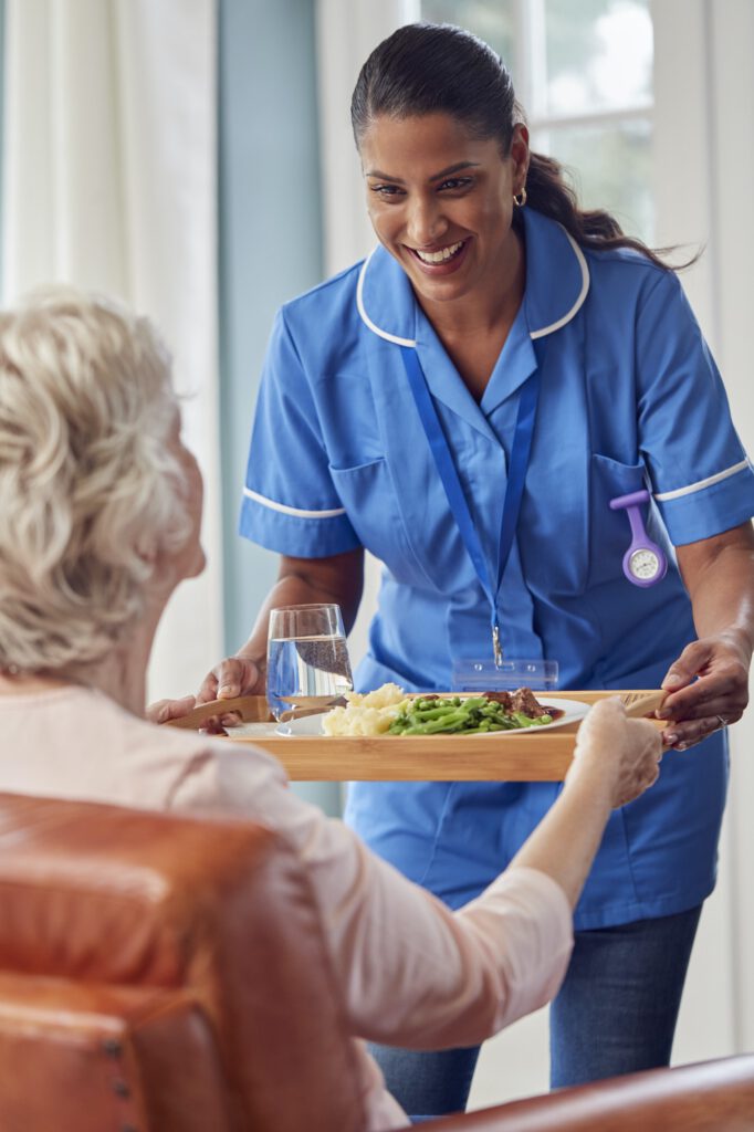 Female Care Worker In Uniform Bringing Meal On Tray To Senior Woman Sitting In Lounge At Home